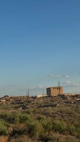 The media file shows a wide, panoramic view of an arid landscape under a clear blue sky with a few scattered clouds. In the mid-ground, an unfinished, multi-story brick building stands prominently, its windows appearing as dark, empty frames. Several tall electrical pylons are visible across the scene, indicating the presence of power infrastructure. The terrain is rough and uneven, covered with dry brush, sparse vegetation, and piles of dirt or debris, suggesting a construction or industrial area, or perhaps an area undergoing development or remediation.

The overall impression is one of a desolate, somewhat remote location. The lighting suggests it is either early morning or late afternoon, with the sun casting long shadows and illuminating the scene with a warm, natural light. The weather appears to be fair and clear. The setting is likely rural or industrial, given the building and infrastructure. There are no people visible in the frame, and no visible text. The emotion evoked is one of quiet stillness and perhaps a sense of abandonment or progress, depending on the interpretation of the unfinished building and the surrounding landscape. The location context provided, Töretam, Kazakhstan, suggests a setting in Central Asia, consistent with the arid environment and the style of the electrical pylons.