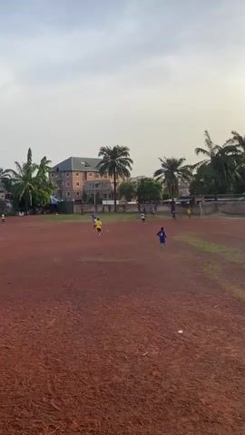 The video shows a soccer game being played on a dirt field, with a few palm trees around the field. There are a few buildings visible in the background. The video then pans to a group of people watching the game from under a covered structure. The spectators are standing and watching the game, with some holding drinks.