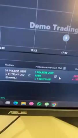 The video shows a computer screen displaying a Demo Trading platform. The screen shows a real-time graph of cryptocurrency prices with time markers. Below the graph are figures labeled "Margin," "Unrealized PNL," and "Realized PNL," all displayed in USDT. The Unrealized PNL fluctuates between approximately 2100 and 2300 USDT, with percentages shown beside it. The computer is an Asus brand, and several app icons are visible on the taskbar including WhatsApp, Telegram, and Google Chrome. The table beneath the monitor has a light tan color.