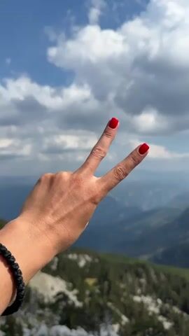 The video opens with a close-up of a person's hand with bright red fingernails, forming a peace sign against a backdrop of a blue sky with white clouds. The camera then pans out to reveal a breathtaking mountainous landscape. The foreground shows a rocky terrain with sparse green trees and dead branches. Further into the distance, layers of mountains recede into a hazy blue, creating a sense of depth. The sky is predominantly blue with scattered, fluffy white clouds, some of which are quite large and casting shadows. The overall impression is of a scenic vista from a high vantage point.