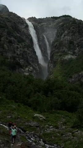 The video shows a tall waterfall cascading down a rocky, forested mountainside under a cloudy sky. Several people are hiking on a dirt path that winds its way up the slope towards the waterfall. The path is rocky and uneven, with patches of snow visible. The hikers are spread out, with some closer to the camera and others further up the path. The surrounding vegetation is dense and green, with trees and bushes covering the lower slopes of the mountain. The waterfall appears to be split into two sections, with water flowing down both. The mist from the waterfall creates a hazy effect in the air.