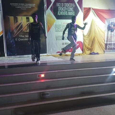 Here is a detailed description of the video:

The video shows two people dancing on a small stage in front of two large banners. The banners are advertising different events, including the "Face of Okokrocity & Okokrocity Iconic Achievers Award" and "The King & Queens." One dancer is wearing a purple jersey with the number 34 and grey pants, while the other is wearing a black jersey with the number 24 and black pants with white stripes. They are performing synchronized dance moves, seemingly to a beat that is not clear in the recording. The stage is next to a set of stairs that lead up to what seems to be a hallway. The setting appears to be an event venue. The video is shot from a low angle and is oriented sideways.
