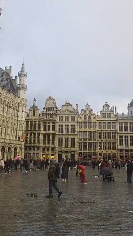 The video shows a square filled with people, likely tourists, in front of a large, ornate, Gothic-style building. The ground is covered with cobblestones and appears wet. The sky is overcast. People are walking around, some are taking pictures, and others are simply looking at the architecture. A vintage-style car is parked near the building. The overall atmosphere seems calm and observant.