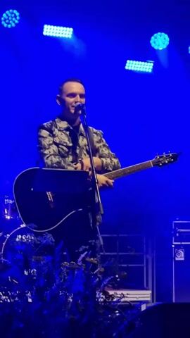 A man with a shaved head and a patterned jacket stands on a stage, holding an acoustic guitar and a microphone. He is singing or speaking into the microphone. Behind him, blue stage lights illuminate the dark background. To his left, a drum set is partially visible. In front of the man, there are some dark, leafy plants. The man is looking towards the right of the frame.