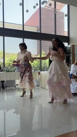 Two women are dancing in a brightly lit indoor space with large windows. The woman on the left wears a light-colored, ruffled blouse and a floral skirt, paired with high heels. She has short, dark hair and is smiling. The woman on the right wears a strapless, tiered white dress with pink floral accents and high heels. She has long, dark hair and is also smiling. They are holding hands and appear to be enjoying themselves, possibly at a celebratory event like a wedding or party.

The setting features a modern interior with a polished floor and high ceilings. Large windows offer a view of lush green trees and buildings outside, suggesting it is daytime and the weather is pleasant. Tables draped with white cloths and decorated with floral arrangements are visible in the background, hinting at a catered event. The overall atmosphere seems joyful and elegant. There is no visible text in the media file.