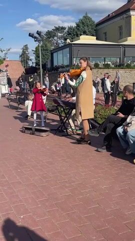 Here is a description of the video:

The video shows a group of musicians performing on a brick-paved public area. A young girl in a red coat stands on her violin case, playing alongside a woman in a tan coat who is also playing a violin. A keyboard player is seated behind them, providing accompaniment. A sheet music stand sits in front of them. Several people are watching the performance, sitting on benches or standing nearby.  In the background, there is a building with large windows and a glimpse of a blue sky with scattered clouds. A light pole stands to the left of the musicians.