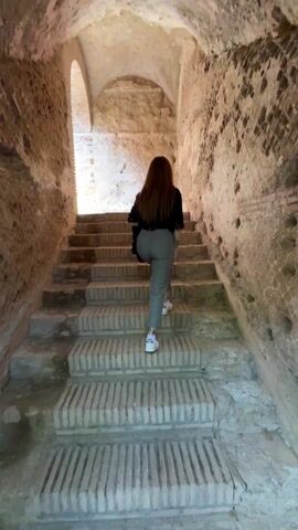 A low-angle shot shows a woman with long red hair climbing a set of stone stairs in an arched tunnel. She is wearing a black shirt, gray pants, and white sneakers, and carries a black bag. The tunnel is constructed of aged stone and brick, with sunlight visible at the top of the stairs. As the woman ascends, two people, a man and a woman, appear in the light at the top of the stairs. The man is wearing a white shirt, while the woman has a camera and is taking photos. The camera is focused on the woman as she walks up the stairs.
