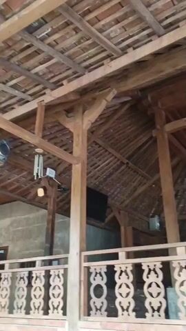 The video pans from right to left, showing the interior of a wooden structure with a tiled roof. The structure has exposed wooden beams and posts, and a decorative wooden railing runs across the foreground. In the background, there are glimpses of the outside, with several motorcycles parked and some trees. There are no people clearly visible in the frame. The overall impression is of an open-air, possibly covered, space with a rustic, wooden aesthetic.