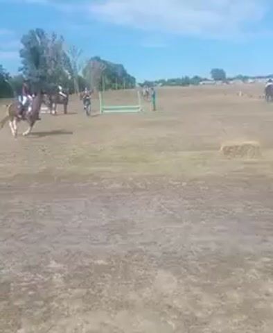 The video shows a horseback riding event in a grassy field under a partly cloudy sky. In the foreground, the ground is dry and patchy. In the middle ground, a brown and white horse is being ridden by a person wearing a white helmet and a red top. The horse is trotting. There is a jump in the background to the left, and two people are standing near it. To the right, there is a bale of hay. In the distance, there are trees, buildings, and more people on horseback. The horse and rider in the middle ground move from left to right across the screen.