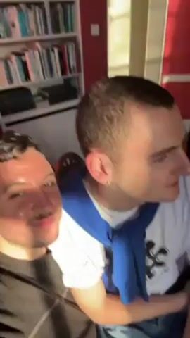Two men are in a room with red walls. One man wears a dark gray sweater and has a mustache. The other man wears a white t-shirt with a design on it and a blue sweater tied around his shoulders. They both have earrings. There is a bookcase filled with books in the background. There is a table with a cup, a newspaper, and bottles of water on it. The men are talking, and the man in the gray sweater smiles at the camera.