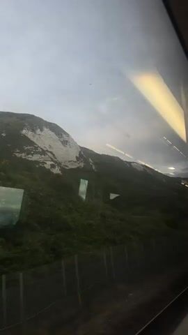 The video shows a view from a train window of a green, tree-covered hillside with a white chalk cliff face on top. The sky is overcast and the light is dim. Reflections of lights from inside the train are visible on the window. A fence runs along the bottom of the hillside. The train appears to be moving.