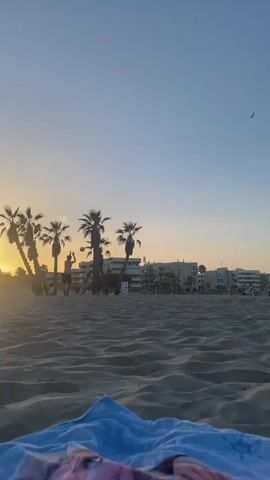 This is a vertical video capturing a serene beach scene at dusk in Canet d'en Berenguer, Spain. The foreground is dominated by rippled sand, with a blue towel draped over the very bottom edge. In the mid-ground, a group of palm trees stands silhouetted against the sky, with a few people milling about among them and on the beach. The sun is setting, casting a warm, golden glow and creating lens flare effects. Beyond the palm trees, buildings line the horizon, suggesting a coastal town. The sky is a clear, gradient of pale blue and soft orange, indicative of sunset. The overall mood is peaceful and relaxing, evoking a sense of end-of-day tranquility on a Spanish beach. There is no visible text in the video.