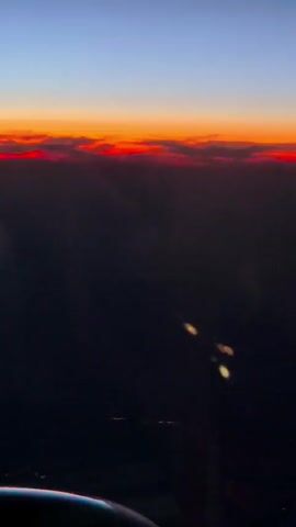 The video shows a view from an airplane window at dusk or dawn. The sky is a gradient of colors, with deep blue at the top fading to orange and red near the horizon. A band of clouds is illuminated by the setting or rising sun, appearing bright red. Below the horizon, the land is dark, with faint lights visible, possibly indicating a city or town. The wing of the airplane is visible in the lower left corner of the frame.