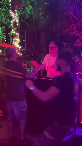 The video shows a DJ at a console, illuminated by purple and pink lights, with two people standing in front of him. The DJ is wearing glasses and a white t-shirt and appears to be mixing music. One of the people in front of him is wearing a black t-shirt with a circular logo and the words "RADIO CAFFE" printed on it. The environment appears to be outdoors, with trees and foliage visible in the background, decorated with string lights.
