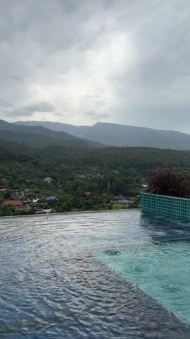 The video shows a view from an infinity pool looking out over a lush green valley and mountains under a cloudy sky. The water in the pool is rippling, and there are some bubbles visible. In the distance, there are small houses and some vegetation. The overall atmosphere is serene and natural.