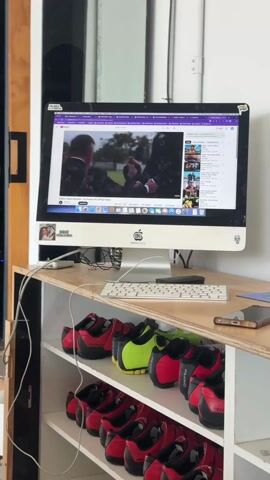 A video shows an office workstation with an Apple iMac desktop computer displaying a wedding video on YouTube. The video on the screen shows scenes from the wedding including the bride and groom exchanging vows, the wedding party, and the bride with her parents. Underneath the desk is a wooden shelf filled with multiple pairs of matching Specialized cycling shoes in black and red, with one pair in black and neon green. A white keyboard and a black mouse are positioned on the desk in front of the iMac, along with a black smartphone. The surrounding environment includes a black doorway and a white wall. The video appears to be shot indoors, likely in a casual office or storage area. A sticker reading "SIGUE PEDALEANDO" is visible on the left side of the computer.