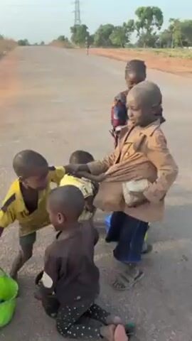 The video shows four young children on a paved road. One child in a yellow shirt and another in a patterned shirt are playfully pushing and interacting with a third child, causing him to fall and spill the contents of a white bucket. The spilled items appear to be small, colorful objects. The fourth child is sitting on the ground near a blue bucket, partially out of view. The children are dressed in worn clothing. The road appears to be in a rural area with trees and a power line visible in the background.