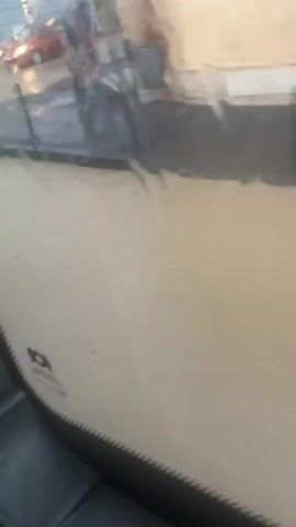 The video shows a street scene during what appears to be heavy rain or a flood. The ground is covered in a significant amount of water, creating reflections of lights from vehicles and buildings. Two red cars are visible, one moving and one stationary. A person, blurred due to motion, is also present in the scene, possibly near a vehicle. The water level suggests that normal street conditions are disrupted. The overall impression is one of an urban environment experiencing adverse weather.