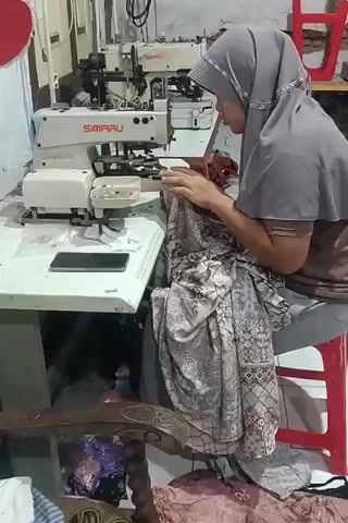 A woman wearing a gray hijab and a brown shirt is seated on a red stool in front of a white sewing machine. She is actively sewing a patterned fabric. The sewing machine is labeled "SIMARU." In the background, another industrial sewing machine is visible. A smartphone is placed on the table next to the sewing machine. The environment appears to be a workshop or tailoring space with various fabrics and sewing equipment.