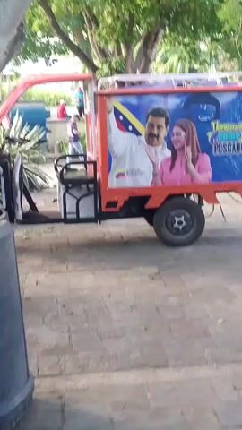 A three-wheeled vehicle with an orange frame and a white cargo bed is parked on a paved surface. The cargo bed is decorated with a large graphic featuring two individuals, a man and a woman, against a background that includes the Venezuelan flag. The man is wearing a white shirt, and the woman is wearing a pink jacket. Trees and foliage are visible in the background, suggesting an outdoor setting.