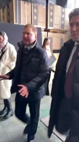 Here's a description of the video:

The video shows a group of people, mostly men in winter coats, walking through a large warehouse-type space. There are stacks of boxes and pallets, suggesting a storage or distribution facility. The people appear to be a delegation, possibly touring the facility. One man in a suit and tie is notably smiling and seems to be the focus of attention. 

As they walk, the camera moves to show flags (Ukraine, the European Union, Italy), and a table set up with microphones, suggesting a press conference or event might be taking place. Overall, the scene appears to be a visit to a warehouse or distribution center, potentially by dignitaries or officials.