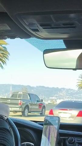 A first-person view from inside a vehicle shows a black pickup truck driving on a highway next to other vehicles. A palm tree is visible outside the window. The sky is clear and blue, and mountains are in the distance. The interior of the vehicle includes a steering wheel and a smartphone mounted on the dashboard displaying a navigation app. The scene appears to be during the daytime.