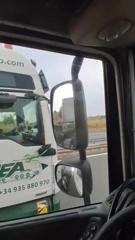 The video is recorded from the driver's perspective inside a truck cab. The driver's hands are visible on the steering wheel. Through the side window, another truck with the logo "ANGEL VELASCO" and "AVEAL" is seen driving alongside. The other truck has a green dumpster attached to its trailer. A GPS device is mounted on the dashboard. The video captures the movement of the trucks on a highway under a cloudy sky.