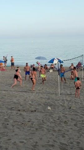 A group of people are playing volleyball on a sandy beach. The ocean is in the background. The sky is a light blue. The people are wearing swimsuits. There is a volleyball net set up. A few people are sitting under umbrellas in the background.