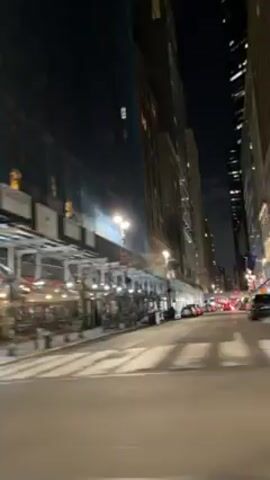 A vehicle moves slowly along a city street at night. Buildings line the street, some under construction with scaffolding. Lights illuminate storefronts. Crosswalks are visible.