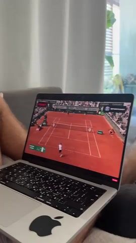 A high-angle, eye-level shot captures a person sitting on a sofa, watching a tennis match on a laptop. The person's bare legs are visible, with the laptop resting on their lap. The tennis match is displayed in a widescreen format, showing a clay court with two players. The laptop has an Apple logo on its base and a black keyboard. The background features light-colored curtains and a window with some greenery visible outside.