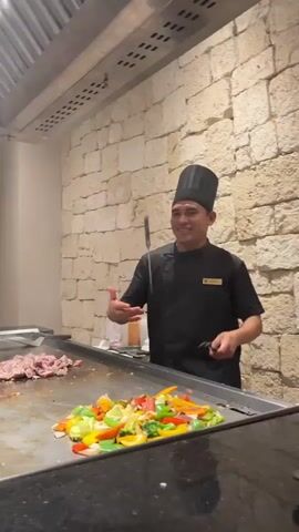 A male chef wearing a black chef's hat and uniform is cooking on a flat-top grill. He is smiling and appears to be interacting with someone off-camera. On the grill, there is sliced meat on the left side and colorful chopped vegetables, including bell peppers, broccoli, and onions, on the right. He is using two spatulas to flip and move the food. Behind the grill is a stone wall and a metal ventilation hood. A woman is partially visible in the background on the left, seated and looking towards the grill.