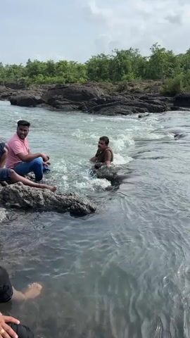 The video shows a group of people by a river. One man in a pink shirt and jeans is sitting on a large rock on the left side of the frame, facing right. Another man in a brown shirt is in the water, appearing to struggle or play in the strong current. His head and shoulders are visible above the water, and he is positioned among dark rocks in the river. Two other people are partially visible in the foreground, one with their legs in the water and the other with their legs crossed. The river has rocky banks and is surrounded by lush green vegetation in the background. The water is turbulent with visible waves and white water.