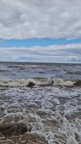 A body of water with waves crashing on a beach. A large rock is visible on the left side of the frame. The sky is cloudy with patches of blue.