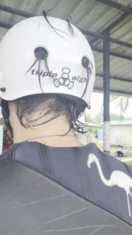 Two individuals are preparing for an activity at an outdoor facility, likely a water sports center, given the presence of wakeboards and helmets. The scene is set under a covered structure with a corrugated metal roof, suggesting an open-air environment. The weather appears overcast, and it might be raining or recently rained, as indicated by the wetness on the ground and the water droplets on the helmet.

The foreground shows the back of a person wearing a white "Triple 8" brand helmet, with strands of dark hair sticking out through the ventilation holes. This person is also wearing a black life vest with a white flamingo design on the back.

In the background, a man in a red t-shirt with yellow and black graphics is standing behind a yellow table. On the table are two wakeboard boots attached to a wakeboard. The man is looking down at the wakeboard, possibly adjusting or preparing it. Behind him, several other wakeboards are propped up. Further in the background, a building with white walls and a dark roof is visible, along with some greenery and dark, round objects that might be tires or buoys.

The overall impression is one of preparation for an athletic activity, possibly wakeboarding or a similar water sport. The overcast weather and indoor/outdoor setting contribute to a functional, sporting atmosphere. The visible text includes "Triple 8" on the helmet, "FOR TEETH" on the wakeboard, and potentially other text on the man's t-shirt which is difficult to discern clearly. The time of day is indeterminate due to the overcast conditions.