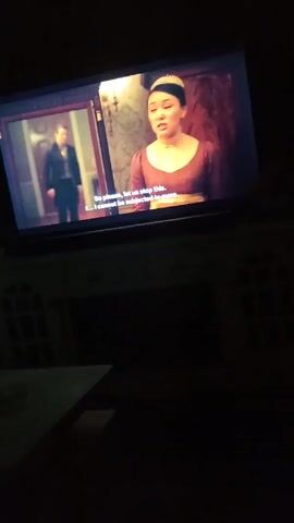 The video depicts a scene from a TV show, captured from a low angle. The screen displays two actors in period costumes within what appears to be an opulent interior. A woman in a brown dress with a white cap pleads, "So please, let us stop this. I cannot be subjected to more..." A man in a dark jacket responds, "Sophie, please, I will leave first, so no servants see me." The room's backdrop features rich mahogany-colored doors and marble accents. The TV is positioned on a white cabinet with decorative arched openings. The surrounding environment is dark, save for the light from the screen. A dark table is visible in the foreground.
