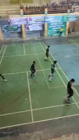 The video shows an indoor sports court with people playing a game. The court is marked with lines for different sports, possibly badminton or volleyball, with a green surface. There are spectators in the bleachers above the court. Several individuals, appearing to be young athletes dressed in athletic wear, are actively engaged in the game. They are spread across the court, moving and interacting with each other, suggesting a competitive match is underway. The lighting is typical of an indoor gymnasium. The scene is viewed from a slightly elevated perspective, looking down onto the court.