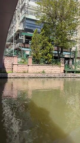 The video depicts a boat ride along a canal in Eskisehir, Turkey, on a clear, sunny day. The initial shots are taken from under a bridge, showing the murky green-brown canal water reflecting the surrounding structures and greenery. A building on the far bank has partially visible text, "BAT...". A young man in a white t-shirt is seen sitting in the boat, looking downwards. Another frame shows a close-up of an orange, fluffy item, likely a blanket or part of the boat's upholstery. The main view is from the front of a dark, gondola-style boat as it moves along the canal. Modern buildings and abundant trees line both sides of the waterway, with another boat visible in the distance, carrying passengers. The sky is bright blue, and the overall scene suggests a leisurely, pleasant activity.