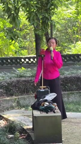 A woman is singing outdoors in what appears to be a park or garden in Banan, China. She stands centered in the frame, wearing a bright pink long-sleeved top and dark wide-legged pants. She holds a green microphone to her mouth, actively singing. To her left, a red smartphone is mounted on a golden tripod stand, positioned next to a portable sound system or speaker setup on a green concrete bench. Various items, including a black bag and what look like sandals, are also on the bench. The background features lush green trees and shrubs, with a traditional-style low wall visible behind them. The scene is brightly lit by daylight, suggesting a clear day. The woman appears focused on her performance.