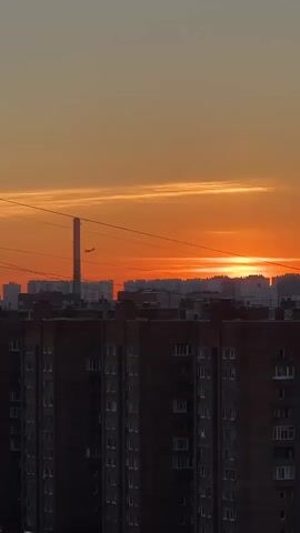 A vertical, low-angle shot captures a city skyline at sunset. The sky is a vibrant orange, with thin, elongated clouds streaking horizontally. In the distance, the setting sun is a bright orange disc peeking over a row of buildings. Closer to the foreground, a tall, cylindrical industrial structure, possibly a smokestack, stands prominently. Below this, the dark silhouettes of several multi-story brick apartment buildings dominate the lower half of the frame. Power lines are visible crisscrossing the sky. The scene is static, with no people or movement evident.