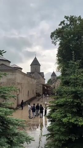 The video shows a group of people standing on a wet, paved pathway in front of a stone building with a domed roof. The sky is overcast, and there are trees on the right side of the frame. The people are casually dressed and appear to be in conversation. The building looks like a historical or religious structure, possibly a monastery or church, with architectural details suggesting it's quite old. The ground is reflecting the light, indicating recent rain.