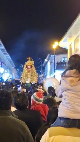 A nighttime outdoor street procession is underway. A large, ornate, gold-colored statue is being carried aloft on a platform by a group of people. The statue appears to be religious in nature, possibly a depiction of a saint or deity, adorned with intricate designs and a crown. A dense crowd of onlookers lines both sides of the street, many of them holding up their phones to capture the event. Smoke or mist is visible in the air around the statue. Streetlights illuminate the scene, casting a warm glow on the buildings and the sky above, which is dark with no visible stars. In the foreground, a young girl with dark hair is perched on someone's shoulders, facing the procession and raising her arms. Several other people are visible in the foreground, their backs to the camera, also looking towards the procession. The architecture of the buildings suggests a historic urban setting, with traditional facades and balconies. The atmosphere appears festive and crowded, indicating a significant cultural or religious gathering.