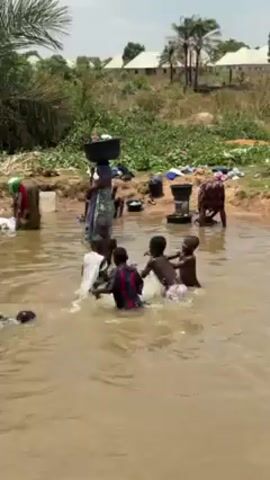 The video depicts a group of people, predominantly women and children, in and around a river or shallow body of water. Several children are actively playing in the water, splashing and interacting with each other. Some are partially clothed, while others are shirtless. On the banks of the water, women are engaged in what appears to be laundry work, using basins and other containers to wash clothes. A woman is balancing a container on her head while walking along the water's edge. The environment includes vegetation, trees, and buildings in the background, suggesting a rural or semi-rural setting. The water appears murky. The scene conveys a sense of daily life and activity centered around the water source.