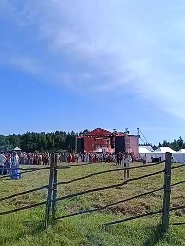 A wide shot captures a large outdoor gathering under a clear blue sky with scattered clouds. In the foreground, a rustic wooden fence runs horizontally across the frame, separating a grassy field from the main event. Beyond the fence, a crowd of people, many dressed in traditional or costume-like attire, are milling about. A prominent red stage with a large screen or backdrop is set up in the middle ground, suggesting a performance or event is taking place or about to begin. Tents are visible on the right side of the stage area. To the left of the stage, a dark, wooden spire-like structure, possibly a church or a historical building, stands tall. The overall atmosphere appears to be that of a festival or cultural event.
