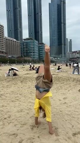 A young woman is performing a handstand on a sandy beach. She is wearing a yellow shirt and brown shorts. Her legs are in the air, and her body is inverted. In the background, there are several tall buildings and a cloudy sky. Other people are visible on the beach, some sitting and others walking. The overall scene is a public beach with a city skyline in the distance.