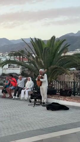 A street musician plays a guitar for a small audience seated on a low wall. The musician is an older man with gray hair and a white shirt and pants. He is standing in front of a microphone and a small amplifier. The audience consists of five people, three men and two women, sitting together on a curved wall. They are all looking at the musician. In the background, there is a large palm tree, buildings, and mountains. The sky is a pale orange and pink, suggesting it is either sunrise or sunset. There is a guitar case on the ground next to the musician. The ground is paved with cobblestones.