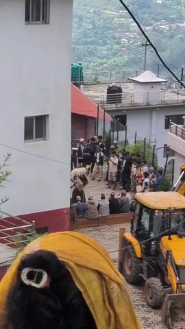 A group of men, some wearing traditional clothing and head coverings, are gathered on a paved street. Several individuals are seated on a low wall. A man in a light-colored garment is interacting with a group of people. In the background, a yellow backhoe is parked. The scene is situated on a hillside with buildings and vegetation visible. A person's head, covered in a yellow fabric with a white object on it, is in the foreground.