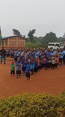 A wide shot captures a large group of young schoolchildren assembled outdoors, predominantly wearing blue uniforms, clapping and singing. They are arranged in rows on a red dirt surface, with a man standing at the front giving direction. Behind the students is a brick building with a sign, a white van with the name "STELLA" on it, and trees. In the foreground, there is a low, green hedge. The sky is overcast. The children are diverse in appearance and size.