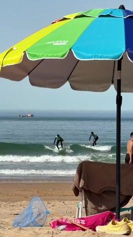 A colorful beach umbrella stands in the foreground, casting a shadow on the sand. Beneath the umbrella, a beach chair is set up, draped with a brown towel. Various beach items, including a pink towel, a blue butterfly net, and a pair of yellow shoes, are scattered on the sand. In the background, two surfers are riding waves in the ocean. One surfer is closer to the shore, while the other is further out. The ocean stretches to the horizon, with a ship visible in the distance. The sky is clear and blue.