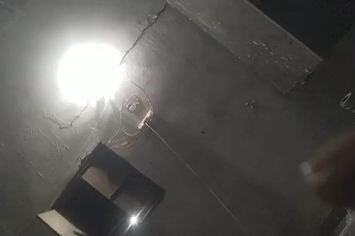 The video displays a dimly lit room, possibly at night. The camera pans across the space, revealing a window with louvers, a standing fan, and a chair. In the foreground, a bed with pillows and a blanket is visible. The walls appear to be made of concrete or a similar rough material. The lighting suggests artificial illumination, possibly from a ceiling light that is intermittently visible. The overall impression is that of a simple, somewhat rustic living space.