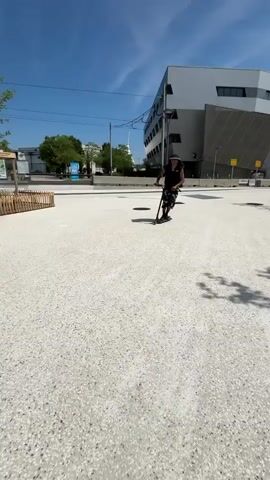 A man performs a scooter trick in an outdoor park. He is Black, wears a black tank top, patterned black and white pants, and a bucket hat. His hair is in dreadlocks. He rides a black scooter on a speckled white concrete surface, approaches a gray rectangular table, jumps the scooter onto the table, balances on a gray rail, and then rides away into the park. The park includes trees, black benches, black trash cans, a wooden fence, and a large white building is in the background, against a blue sky.